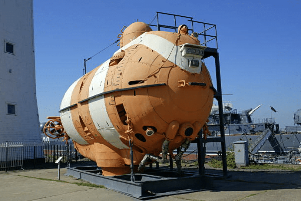 Screenshot 2026-03-16 at 221549 - N9BO℠ | Global Underwater Services Ltd An orange and white deep-sea submersible is displayed outdoors on a metal platform, with a blue sky and other maritime equipment visible in the background.