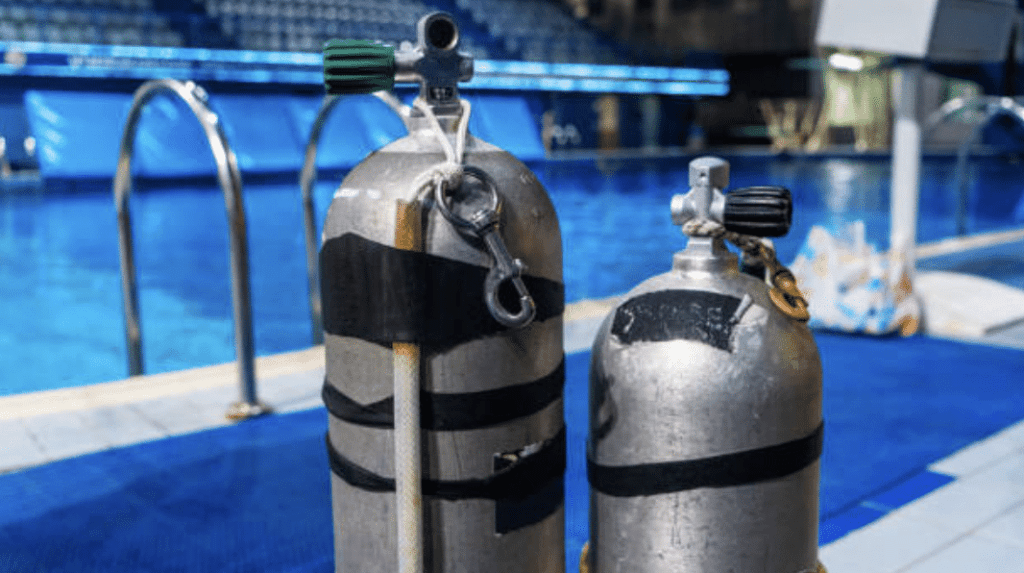 Screenshot 2026-03-16 at 222355 - N9BO℠ | Global Underwater Services Ltd Two scuba diving cylinders stand upright on the edge of an indoor swimming pool, with a metal ladder and diving boards visible in the background.