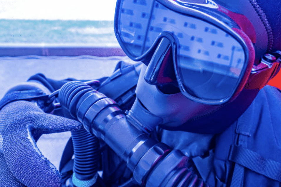 Screenshot 2026-03-16 at 222925 - N9BO℠ | Global Underwater Services Ltd A close-up of a person wearing a black diving mask, gloves, and breathing apparatus, dressed in full scuba diving kit. The reflection in the mask shows a blurred outdoor scene.