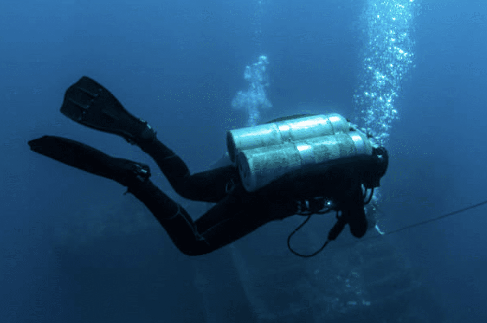 Screenshot 2026-03-16 at 224128 - N9BO℠ | Global Underwater Services Ltd A scuba diver in full kit, including twin oxygen cylinders and fins, swims underwater near a sunken structure, with bubbles rising to the water’s surface.