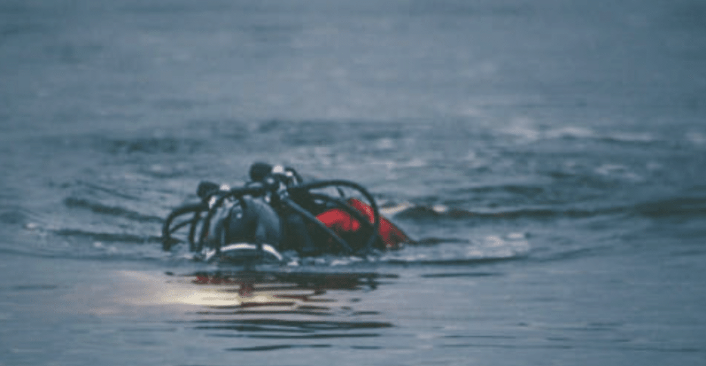 Screenshot 2026-03-16 at 225759 - N9BO℠ | Global Underwater Services Ltd A scuba diver wearing gear and a red cylinder is partially submerged in a calm, blue body of water, with only their head and equipment visible above the surface.