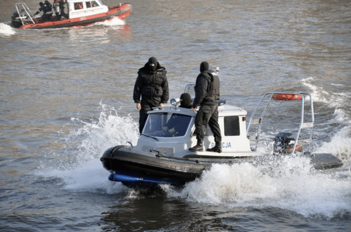 Screenshot 2026-03-16 at 230356 - N9BO℠ | Global Underwater Services Ltd Two police officers in black uniforms ride a small police boat speeding through the water, creating splashes. Another red and white boat with several people is visible in the background.