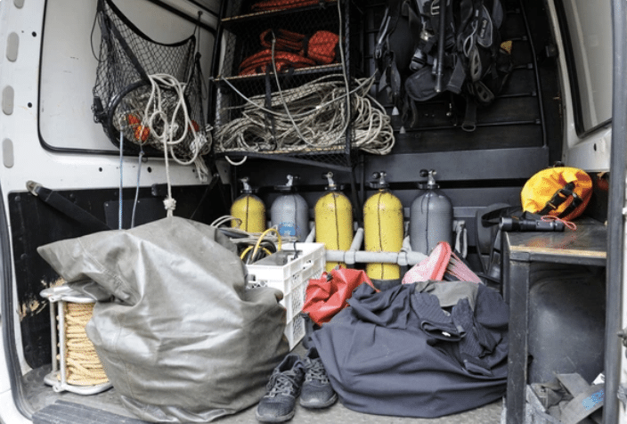 Open van with dive gear inside, including scuba cylinders, ropes, wetsuits, fin bags, a crate, and shoes. Equipment is organised on shelves and the floor, ready for a diving or rescue operation.