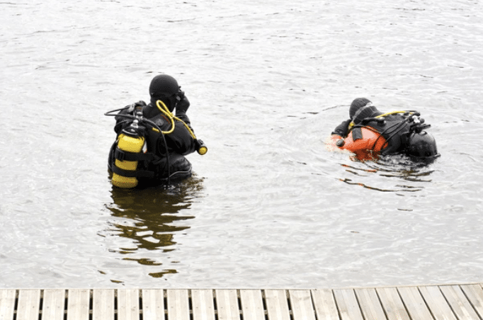 Two scuba divers in wetsuits and cylinders are in the water near a wooden jetty. One diver is standing upright while the other is partially submerged, facing the water.