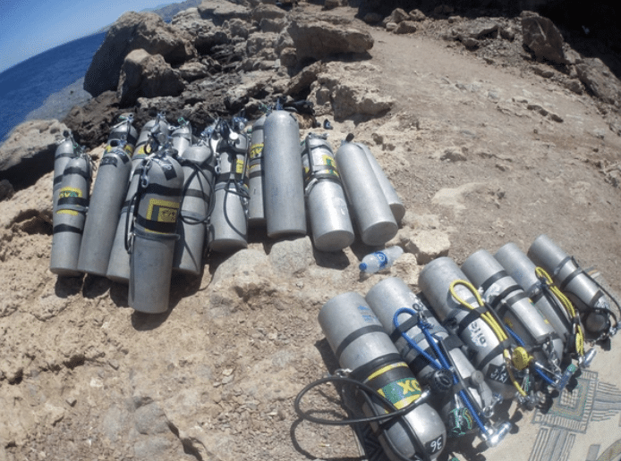 Screenshot 2026-03-16 at 232125 - N9BO℠ | Global Underwater Services Ltd Several scuba diving cylinders and gear are lined up on rocky ground near a coastline, with the sea and rugged rocks visible in the background under daylight.