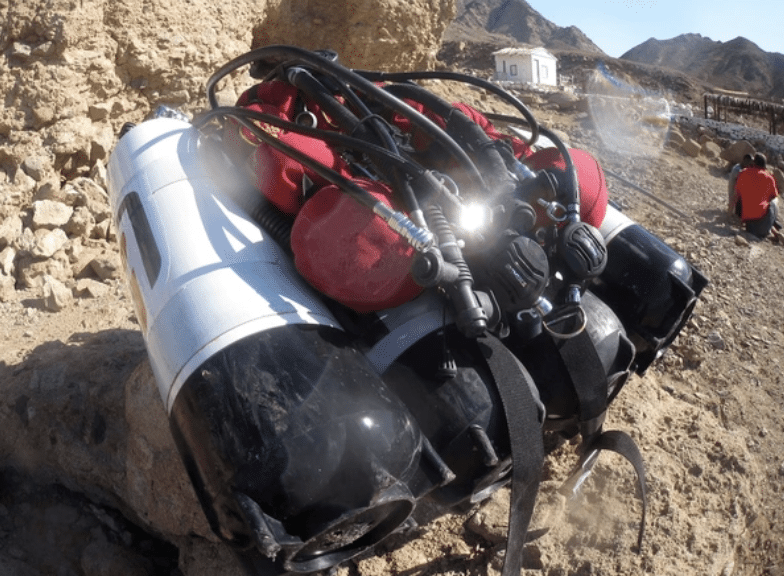 Screenshot 2026-03-16 at 232133 - N9BO℠ | Global Underwater Services Ltd A set of scuba diving cylinders and red buoyancy compensators with attached hoses and regulators rest on rocky ground in a desert-like area, with mountains and buildings in the background.