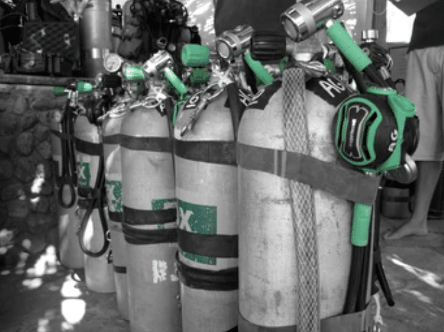 Several scuba diving air cylinders are lined up outdoors. The cylinders are mostly in greyscale except for the vibrant green valves and regulators, which stand out. A person’s legs are visible in the background.