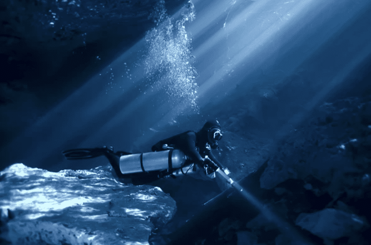 Screenshot 2026-03-16 at 233849 - N9BO℠ | Global Underwater Services Ltd A scuba diver with a torch explores an underwater cave, surrounded by rocks and illuminated by beams of sunlight filtering through the water above.