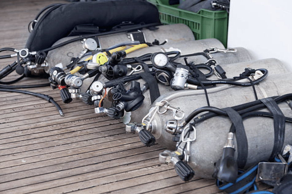 Screenshot 2026-03-16 at 234314 - N9BO℠ | Global Underwater Services Ltd Several scuba diving cylinders with attached regulators and gauges are lined up on a wooden decking, with diving kit and hoses resting on top of them.