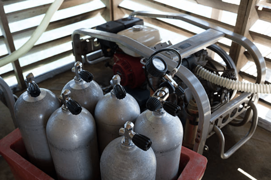 Screenshot 2026-03-16 at 234329 - N9BO℠ | Global Underwater Services Ltd Six silver scuba diving air cylinders with black valves sit in a red container next to a scuba cylinder compressor with a pressure gauge, inside a wooden structure with slatted walls.