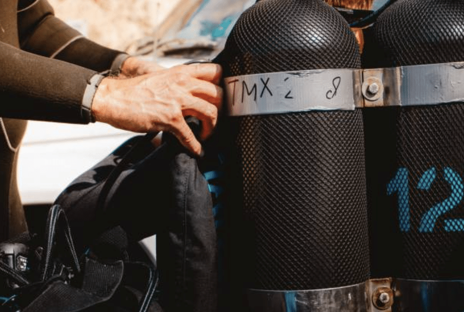 Screenshot 2026-03-16 at 234434 - N9BO℠ | Global Underwater Services Ltd A person in a wetsuit adjusts scuba diving gear, focusing on two large black oxygen cylinders secured with metal straps. The cylinders are labelled with “TMX” and the number 12 in blue.