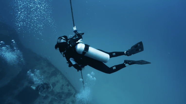Screenshot 2026-03-16 at 235432 - N9BO℠ | Global Underwater Services Ltd A scuba diver in full kit holds onto a rope underwater, surrounded by bubbles, with another diver visible in the background near the seabed.