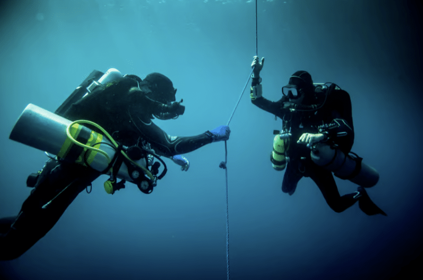 Screenshot 2026-03-16 at 235534 - N9BO℠ | Global Underwater Services Ltd Two scuba divers in wetsuits and gear are underwater, holding onto a rope, surrounded by blue water and illuminated by ambient light from above.