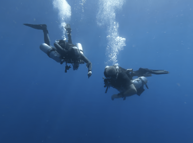 Screenshot 2026-03-16 at 235559 - N9BO℠ | Global Underwater Services Ltd Two scuba divers swim underwater in clear blue sea, surrounded by bubbles, wearing full diving kit including fins, cylinders, and wetsuits.