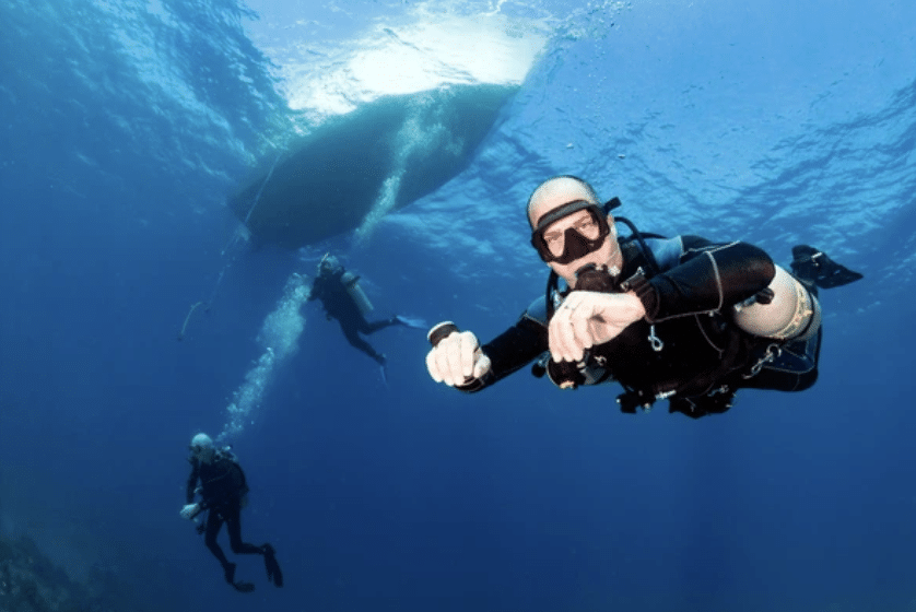 Screenshot 2026-03-17 at 001923 - N9BO℠ | Global Underwater Services Ltd Three scuba divers swim underwater near a boat, with one diver close to the camera giving an OK hand signal, and two others in the background descending towards the sea floor. Sunlight filters through the water’s surface.