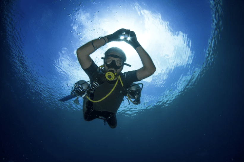 Screenshot 2026-03-17 at 001944 - N9BO℠ | Global Underwater Services Ltd A scuba diver underwater looks up towards the sunlit surface, wearing full diving kit and making a hand signal with both hands, surrounded by deep blue water.