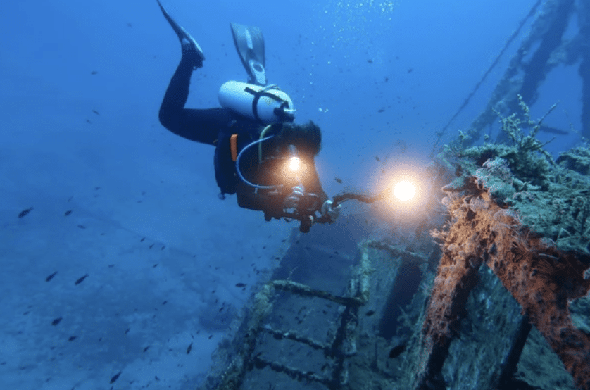 Screenshot 2026-03-17 at 003934 - N9BO℠ | Global Underwater Services Ltd A scuba diver with a torch explores a sunken, rust-covered shipwreck underwater, surrounded by small fish and deep blue water.