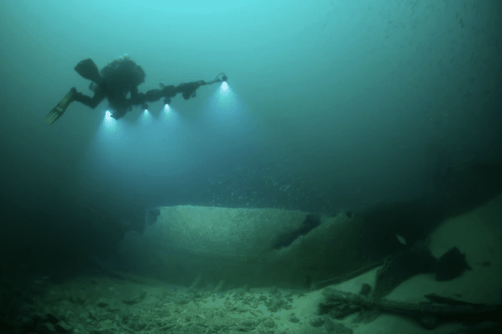 Screenshot 2026-03-17 at 004246 - N9BO℠ | Global Underwater Services Ltd A scuba diver with three bright lights explores the seabed, illuminating the remains of a large underwater shipwreck in murky green-blue water.