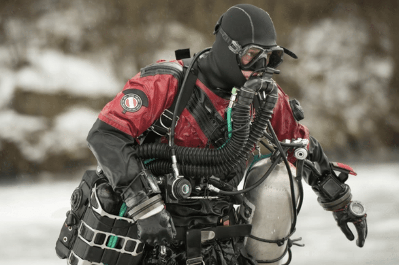 Screenshot 2026-03-17 at 004334 - N9BO℠ | Global Underwater Services Ltd A scuba diver in a red drysuit and black hood wears a full-face mask and rebreather gear, carrying an air cylinder. Snow and ice are visible in the background, indicating a cold environment.