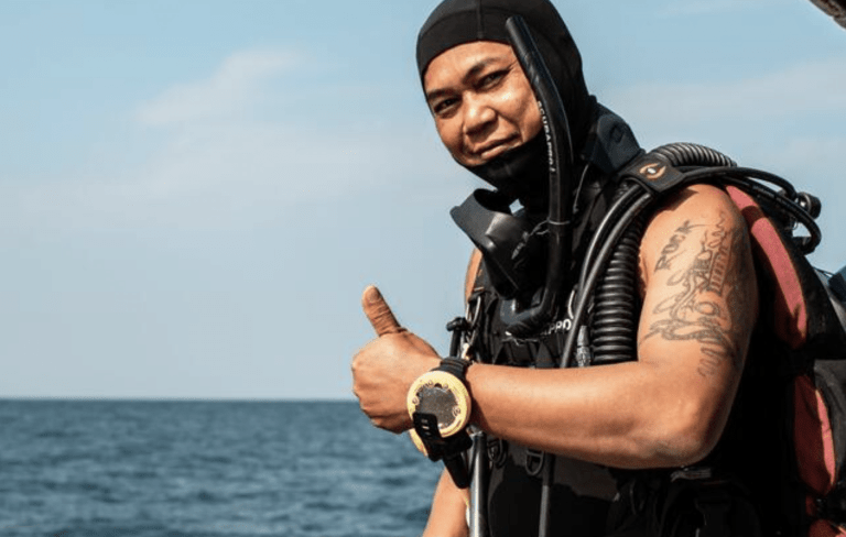 A scuba diver wearing a wetsuit, dive computer, and kit gives a thumbs up near the sea, smiling with a tattooed arm and blue sky in the background.