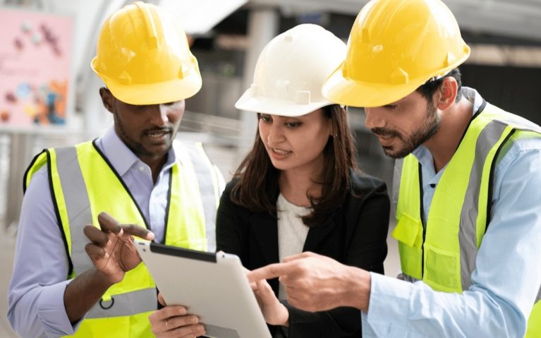 Three construction workers wearing hard hats and high-visibility vests look at and discuss information on a digital tablet, collaborating at an indoor construction site.