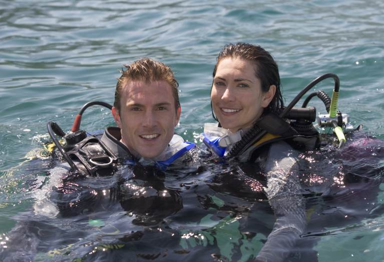 Two people wearing scuba diving gear smile at the camera whilst floating together in clear blue water on a sunny day.