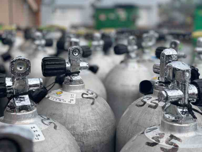 A close-up view of multiple metal oxygen cylinders with black valves, arranged in rows outdoors. The cylinders are labelled with stickers and numbers, and the background is slightly blurred.