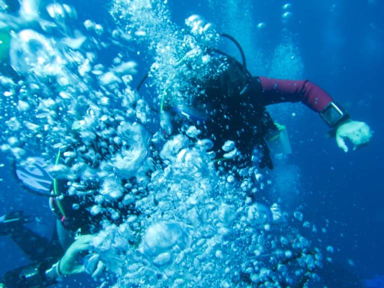 A scuba diver underwater surrounded by a large cloud of rising bubbles, with blue water and diving equipment visible in the background.