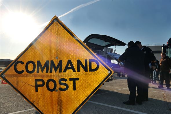A bright yellow diamond-shaped Command Post sign stands in the foreground, with uniformed personnel and an emergency response vehicle visible in the background under a sunny sky.