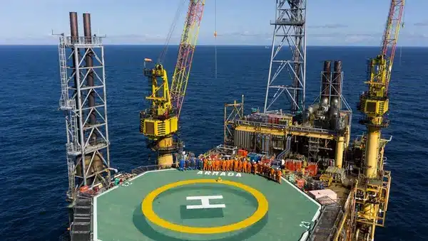A group of workers in orange safety gear stands on a helipad marked ARMADA on an offshore oil platform, surrounded by sea with cranes and industrial equipment visible.