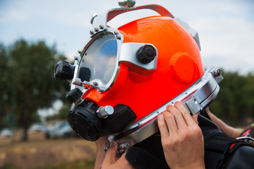 A person adjusts a bright orange deep-sea diving helmet with a transparent visor and metal fittings, preparing for an underwater activity outdoors.