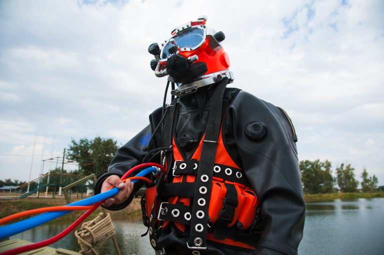 A person wearing a red diving helmet and black wetsuit holds colourful hoses beside a body of water, with trees and a cloudy sky in the background.