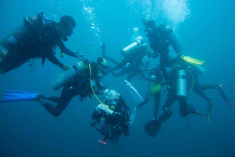 A group of scuba divers underwater, wearing wetsuits and carrying oxygen cylinders, swim closely together in clear blue water. Bubbles rise around them as they communicate and interact.