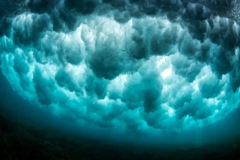 Underwater view looking up at the turbulent, foamy underside of ocean waves, with sunlight filtering through the moving water, creating a dramatic and dynamic scene.