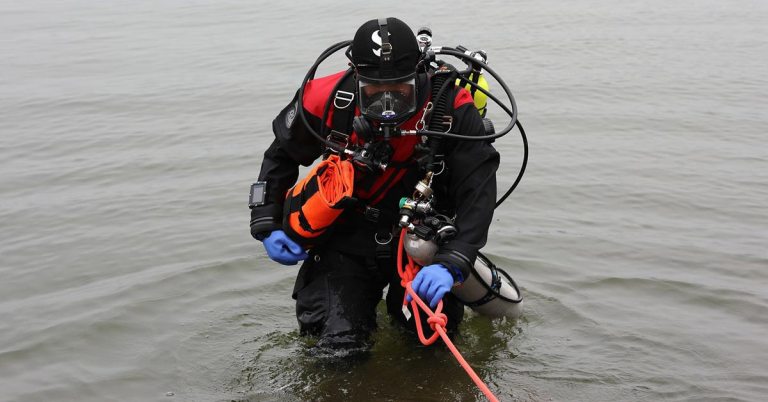 A scuba diver in full kit, including a black wetsuit, gloves, and a helmet, stands waist-deep in water, holding an orange rope and an oxygen cylinder. The background shows a calm body of water.