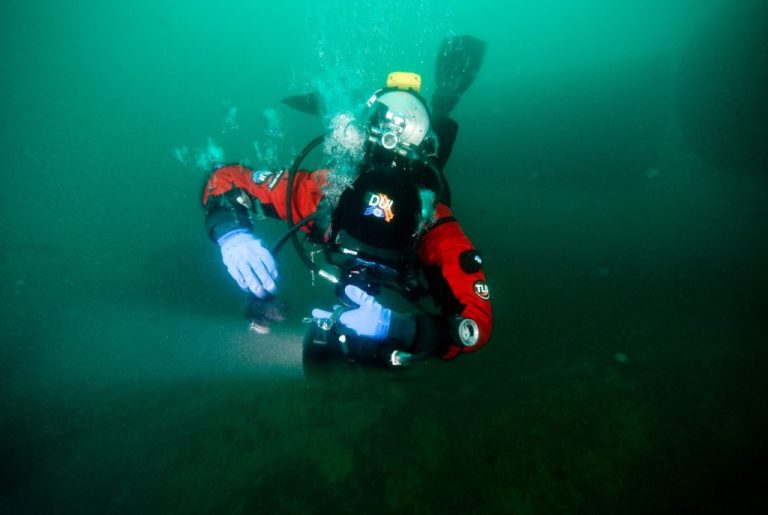 A scuba diver in a red drysuit and helmet examines something on the seabed, shining a torch underwater. Bubbles rise from the diver’s breathing apparatus in the greenish water.