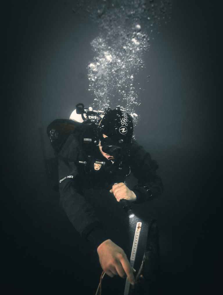 A scuba diver in dark underwater gear holds onto a rope or guide line while ascending or descending in deep, murky water, with bubbles rising above their head.
