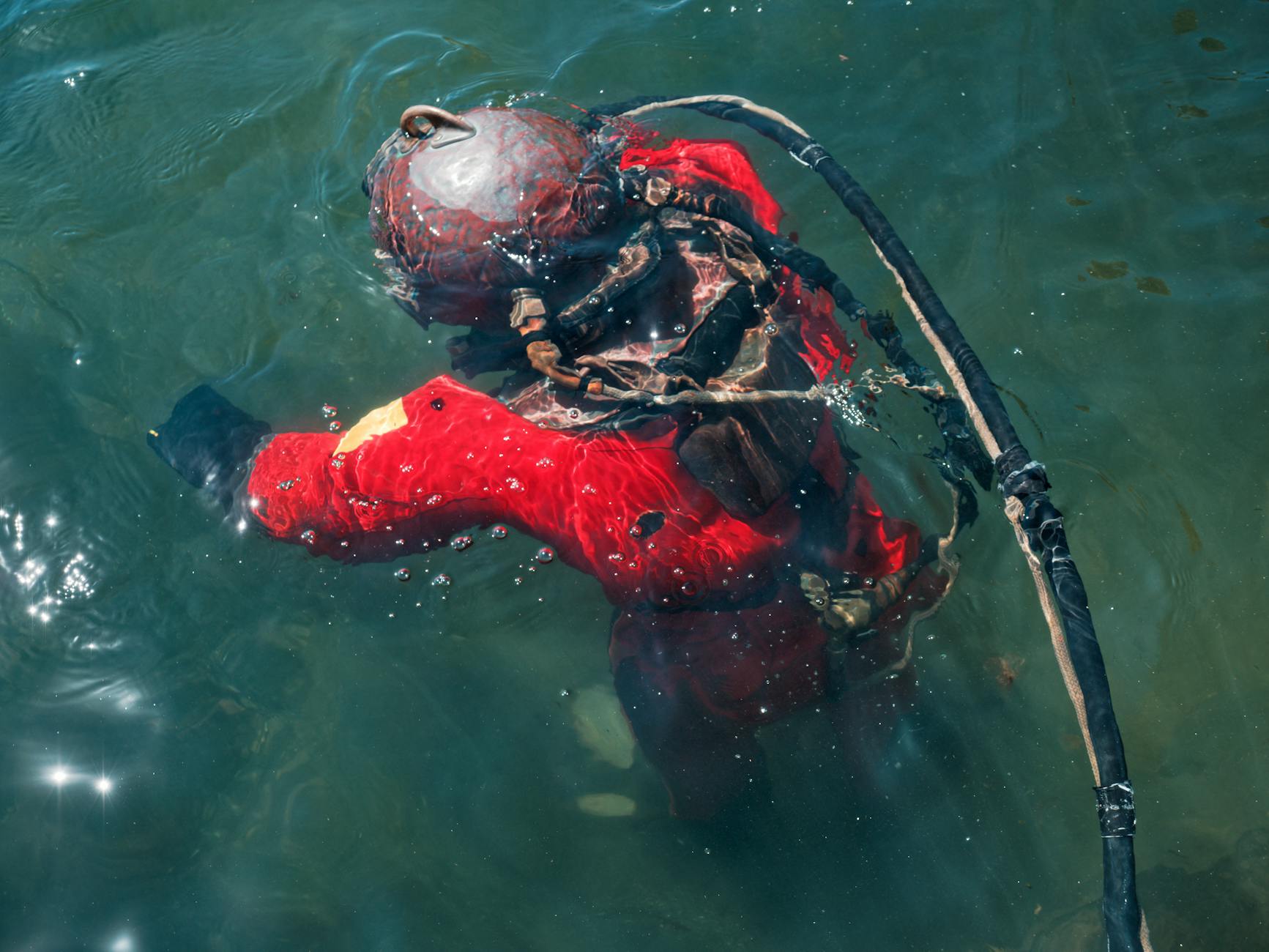 pexels-photo-20481590 - N9BO℠ | Global Underwater Services Ltd A diver wearing an old-fashioned metal helmet and red protective suit is partially submerged in greenish water, holding onto a thick hose or cable connected to their helmet.