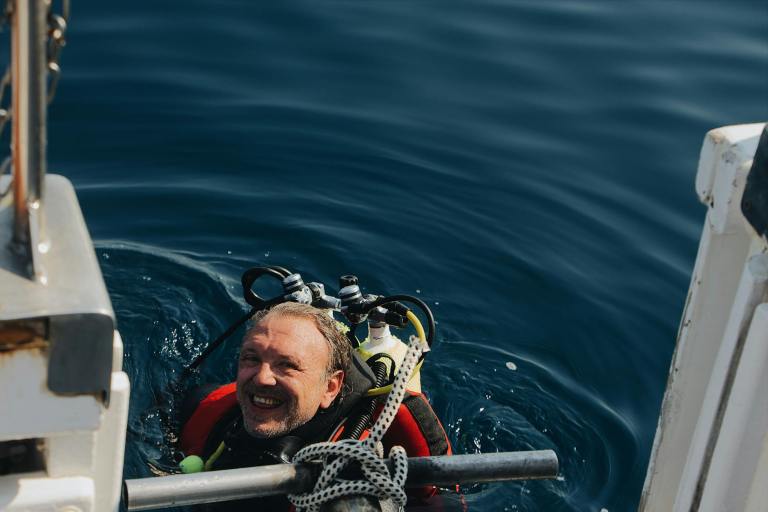 A smiling man in scuba diving gear is in the water next to a boat ladder, preparing to climb aboard on a sunny day.
