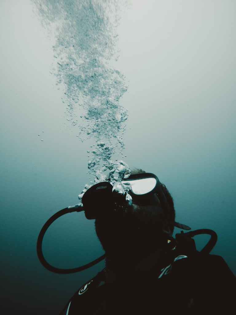 A scuba diver is underwater, exhaling bubbles that rise to the surface. The diver is wearing a mask and breathing apparatus, and the scene has a dark, moody blue tone.