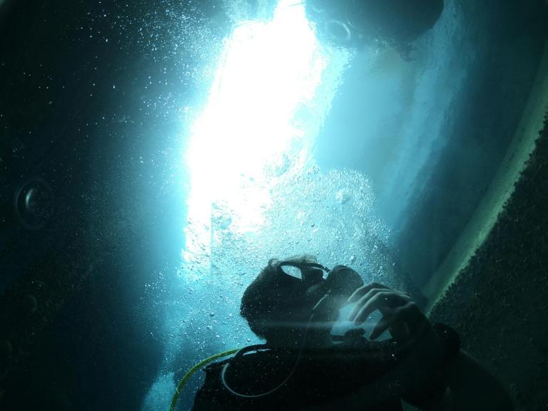 A scuba diver underwater is surrounded by bubbles, with sunlight streaming down from the surface above, creating a bright, glowing effect through the water.