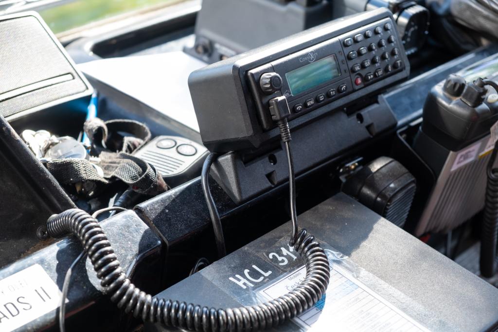 - N9BO℠ | Global Underwater Services Ltd A close-up of a police radio communication device and microphone mounted inside a vehicle, with coiled leads, control buttons, and other equipment visible nearby.