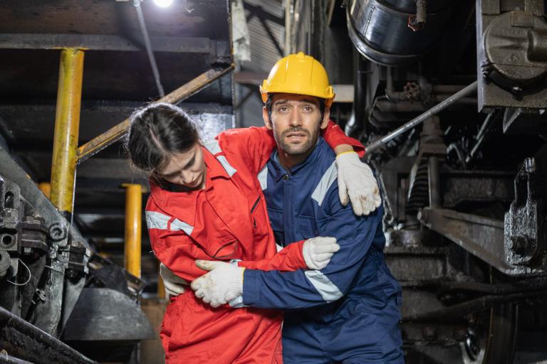 A worker in a yellow hard hat and blue overalls helps an injured colleague in red overalls under industrial machinery, showing concern and support in a factory setting.