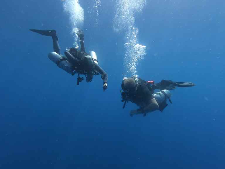 Two scuba divers swim underwater in clear blue sea, surrounded by bubbles. Both are wearing full scuba equipment and fins, moving side by side, appearing to communicate or gesture towards each other.