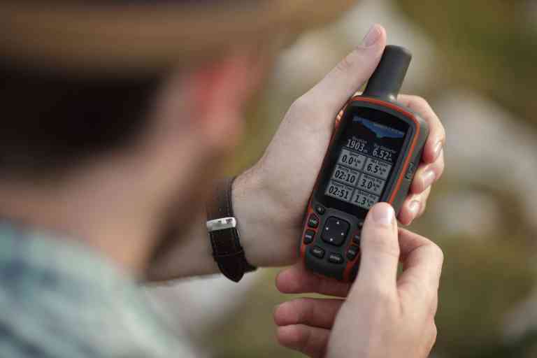 A person wearing a watch and hat holds a handheld satnav device, displaying navigation data on the screen, likely whilst outdoors. The background is blurred.