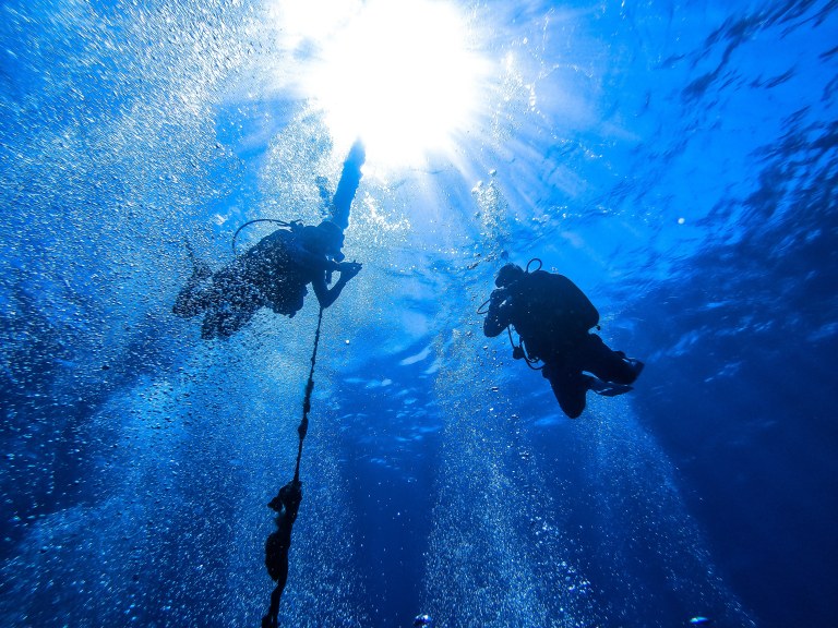 Two scuba divers are underwater, surrounded by bubbles, with sunlight streaming down from the surface above. One diver holds onto a rope while the other swims nearby in the deep blue water.