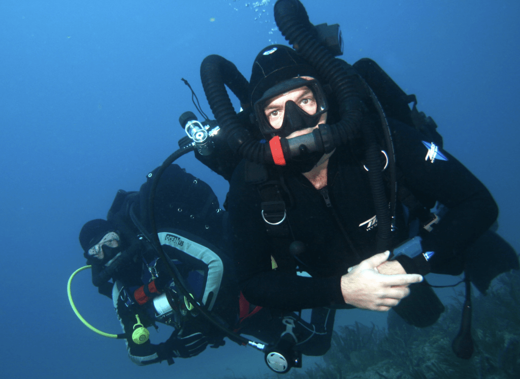 Screenshot 2026-03-30 at 162947 - N9BO℠ | Global Underwater Services Ltd Two scuba divers in black wetsuits and masks swim underwater. One diver is closer to the camera holding equipment, both wearing oxygen cylinders and surrounded by blue water.