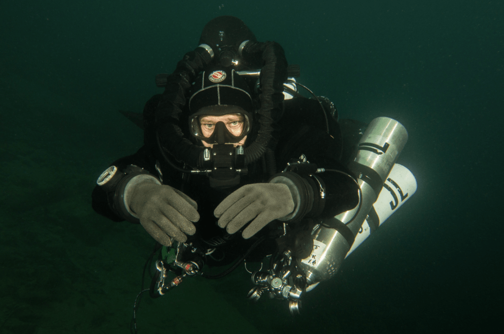 Screenshot 2026-03-30 at 163014 - N9BO℠ | Global Underwater Services Ltd A scuba diver wearing a full-face mask, gloves, and technical diving equipment swims underwater in dark green water while facing the camera, holding their hands out in front.