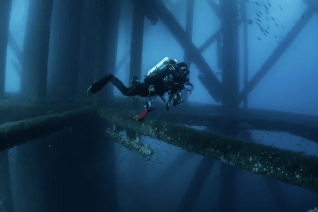 Screenshot 2026-03-30 at 164151 - N9BO℠ | Global Underwater Services Ltd A scuba diver in full kit swims underwater near the metal beams and supports of an offshore oil rig, surrounded by deep blue water and small fish.
