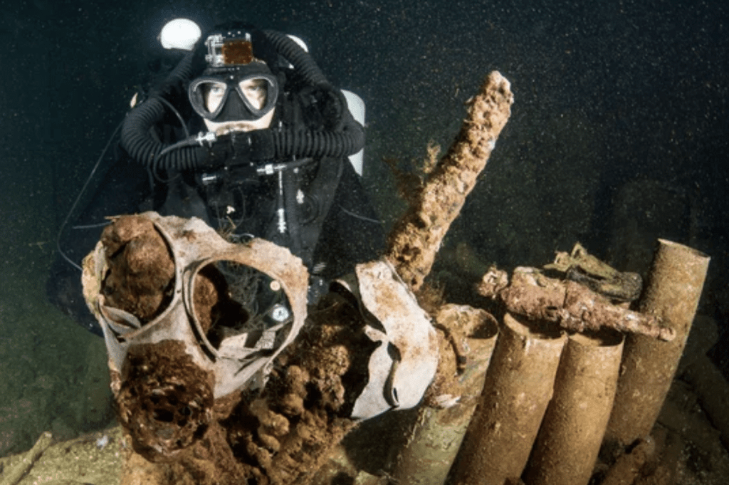 Screenshot 2026-03-30 at 164201 - N9BO℠ | Global Underwater Services Ltd A scuba diver in full kit examines a collection of corroded metal debris and shells underwater, surrounded by dark water and particles.